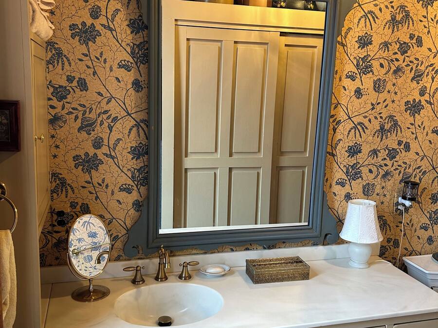 Uma casa de banho com um espelho grande, uma pia, toucador com gavetas, um pequeno banquinho e um painel decorativo com papel de parede floral.