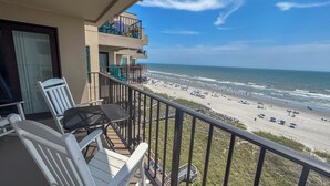 A balcony with two white chairs and a table, overlooking a beach with umbrellas and people. - 20% off week of July 25-Aug. 1.  3BR OF condo. Ocean Drive. Pool. (North Myrtle Beach)