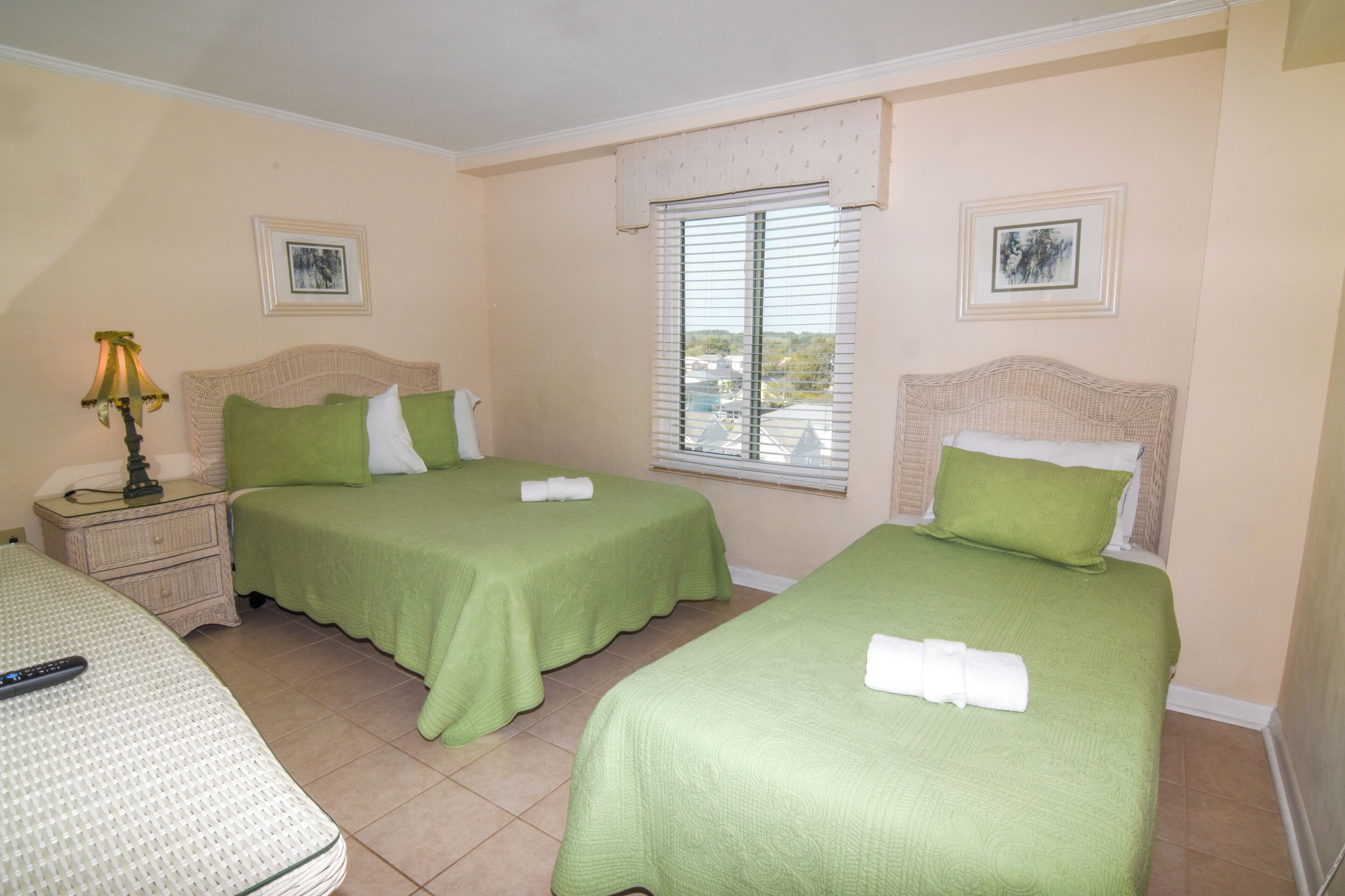 A hotel room with two single beds, a nightstand, a lamp, and a window with blinds.