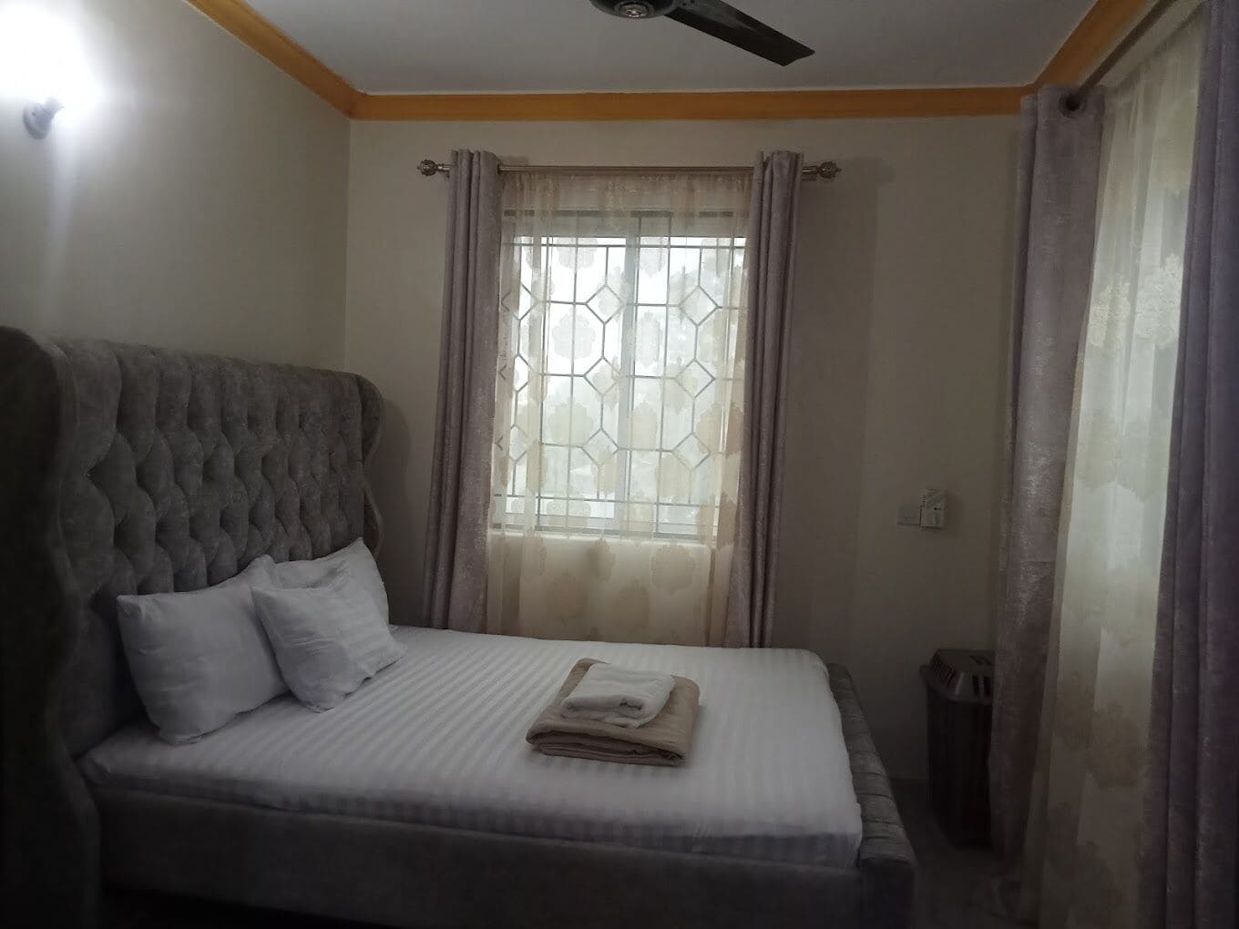 Une chambre à coucher comprenant un lit, un ventilateur de plafond, une fenêtre avec des rideaux et un luminaire fixé au mur.