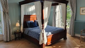 Ein Bett mit Baldachin und Holzkopf, ein Nachttisch mit Lampe sowie Blick ins Freie durch ein Fenster mit Vorhängen.