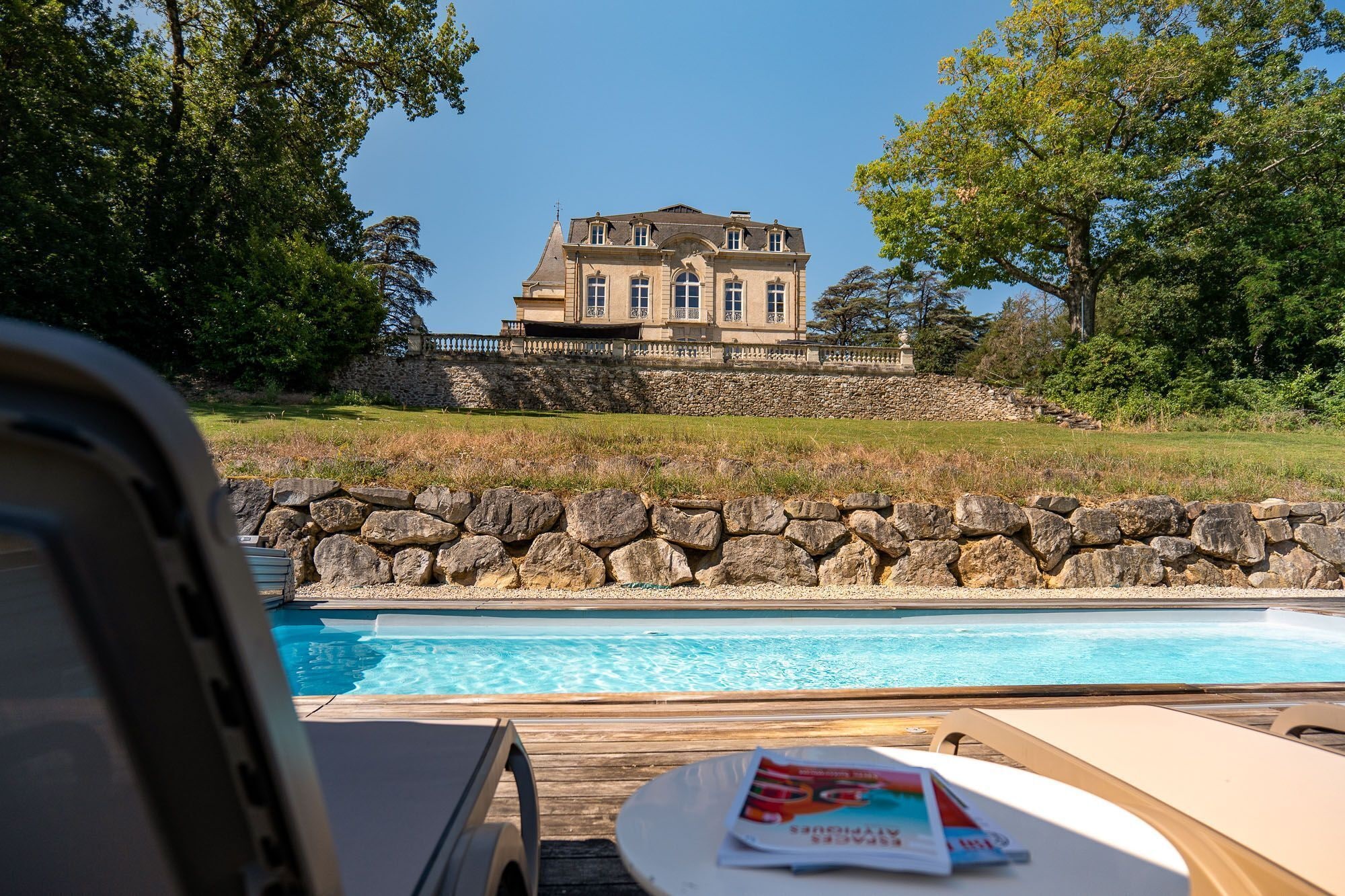 Vue sur une grande maison avec un mur en pierre, prise depuis la piscine, avec un ciel bleu dégagé.