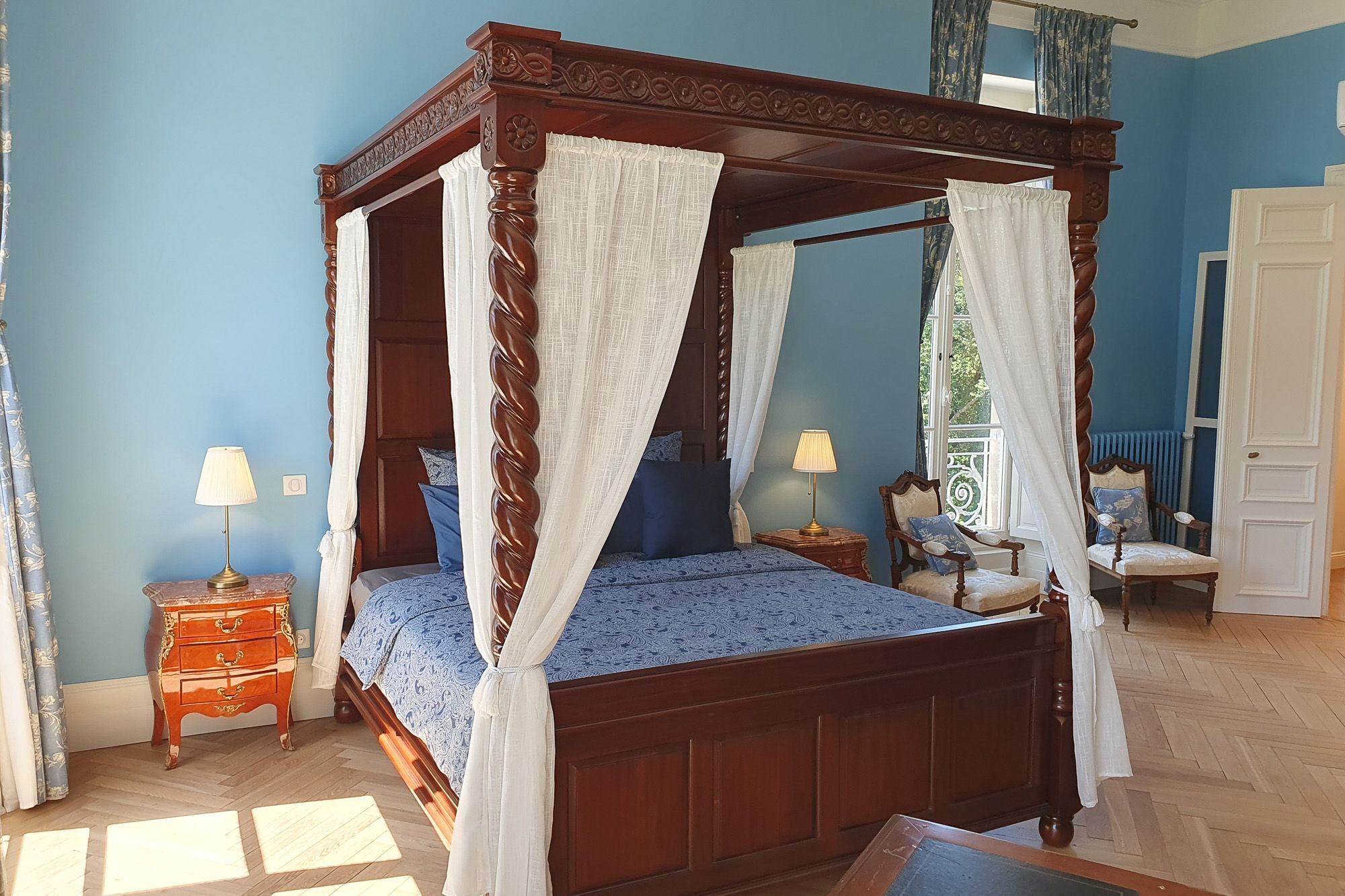 A four-poster canopy bed with a white bedspread, two bedside tables with lamps, and a view of the outdoors through a window.