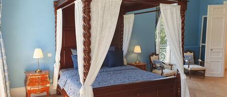 A four-poster canopy bed with a white bedspread, two bedside tables with lamps, and a view of the outdoors through a window.