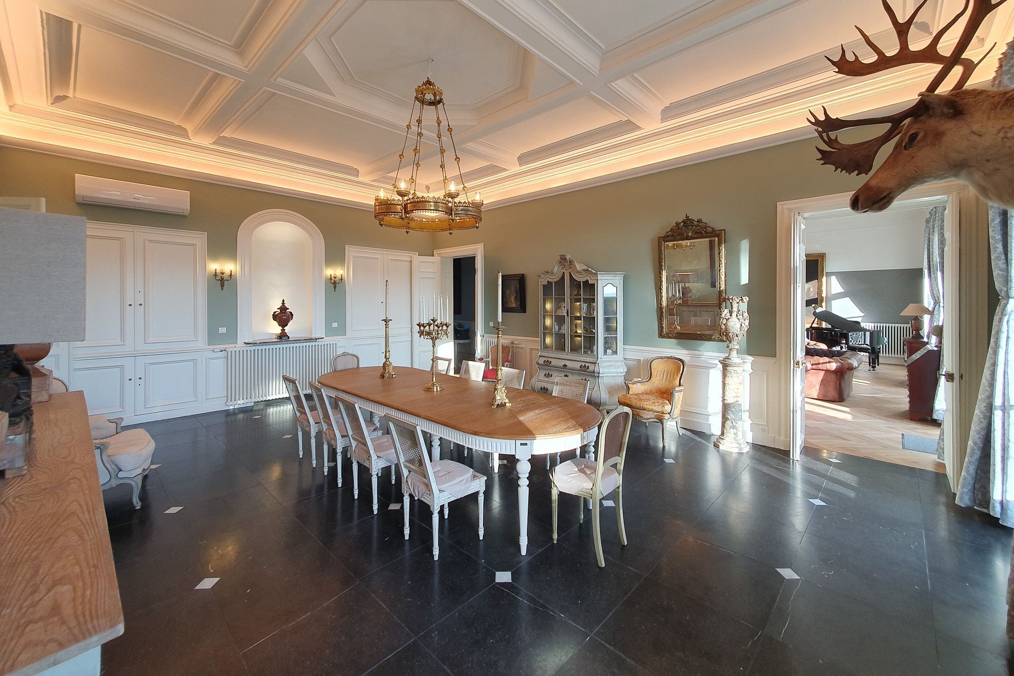 Un vaste espace dédié à la salle à manger, avec une grande table en bois et des chaises à revêtement blanc, un lustre et une tête de cerf fixée au mur.