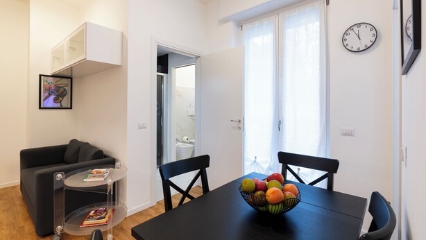 A dining area with a black table, chairs, a fruit bowl, a small round table with a book, a black sofa, and a white cabinet. - notaMI - Civitali Suite - San Siro (Milan)