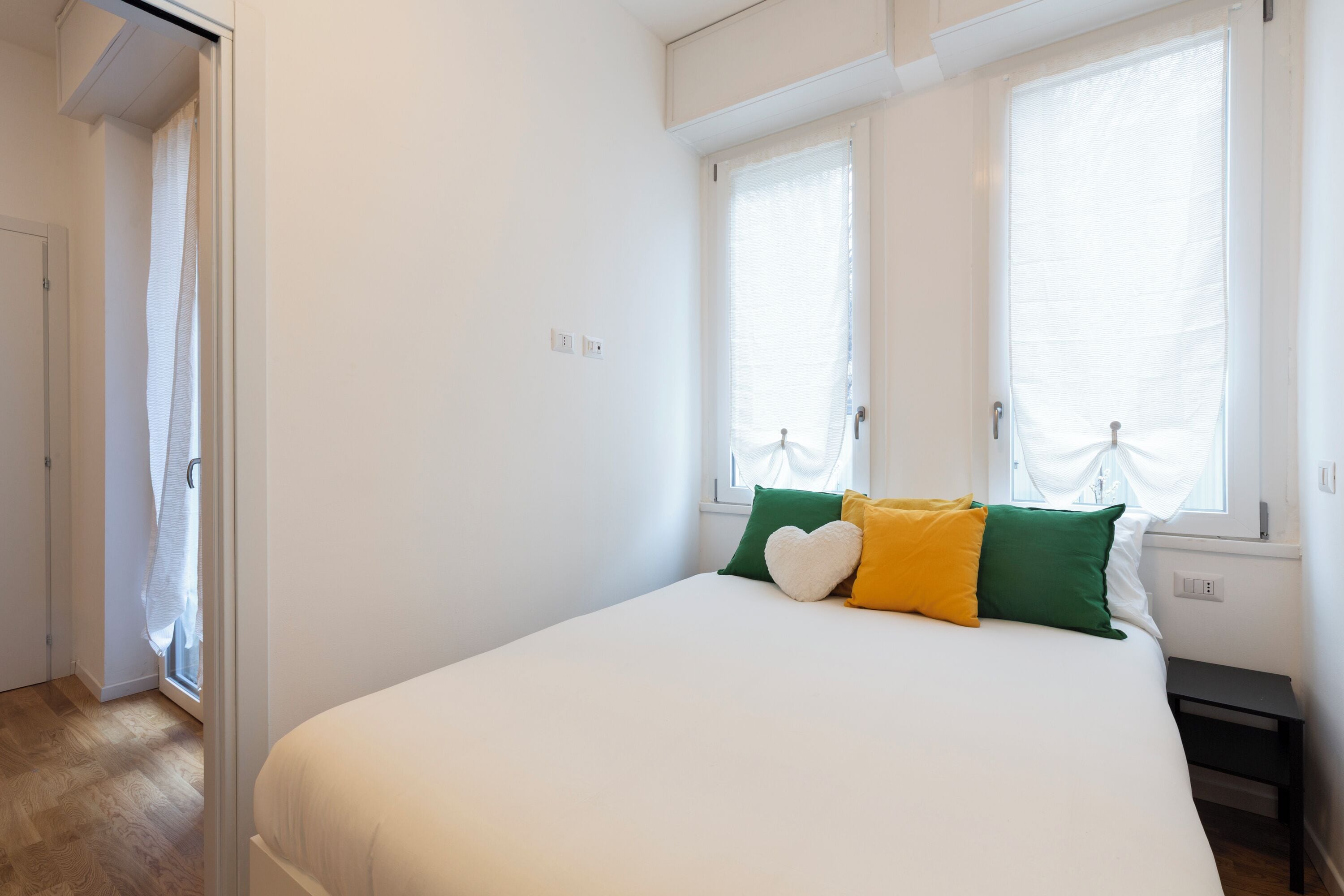 A bedroom with a bed, a window with curtains, a bedside table, and a door leading to another room.