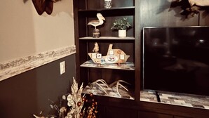 A room with a mounted fish on the wall, a television, shelves with decorative items, and a vase with dried flowers.