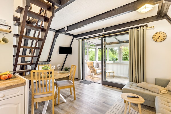 A room with a wooden dining table, chairs, a sofa, a TV mounted on the wall, and a large window with curtains.