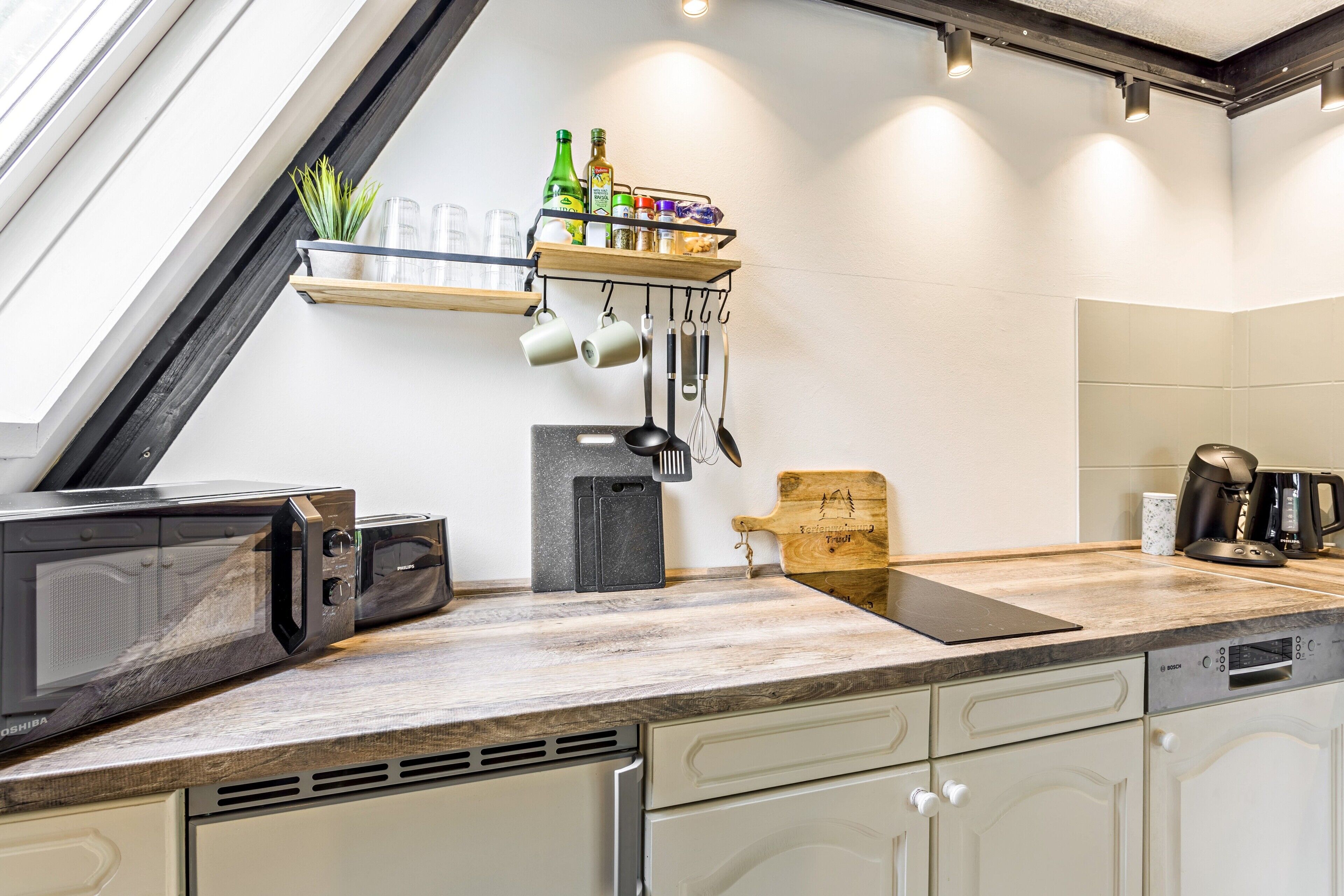 A modern kitchen with a built-in microwave, toaster, and coffee maker. There is a wooden cutting board and a shelf with kitchen utensils and bottles.