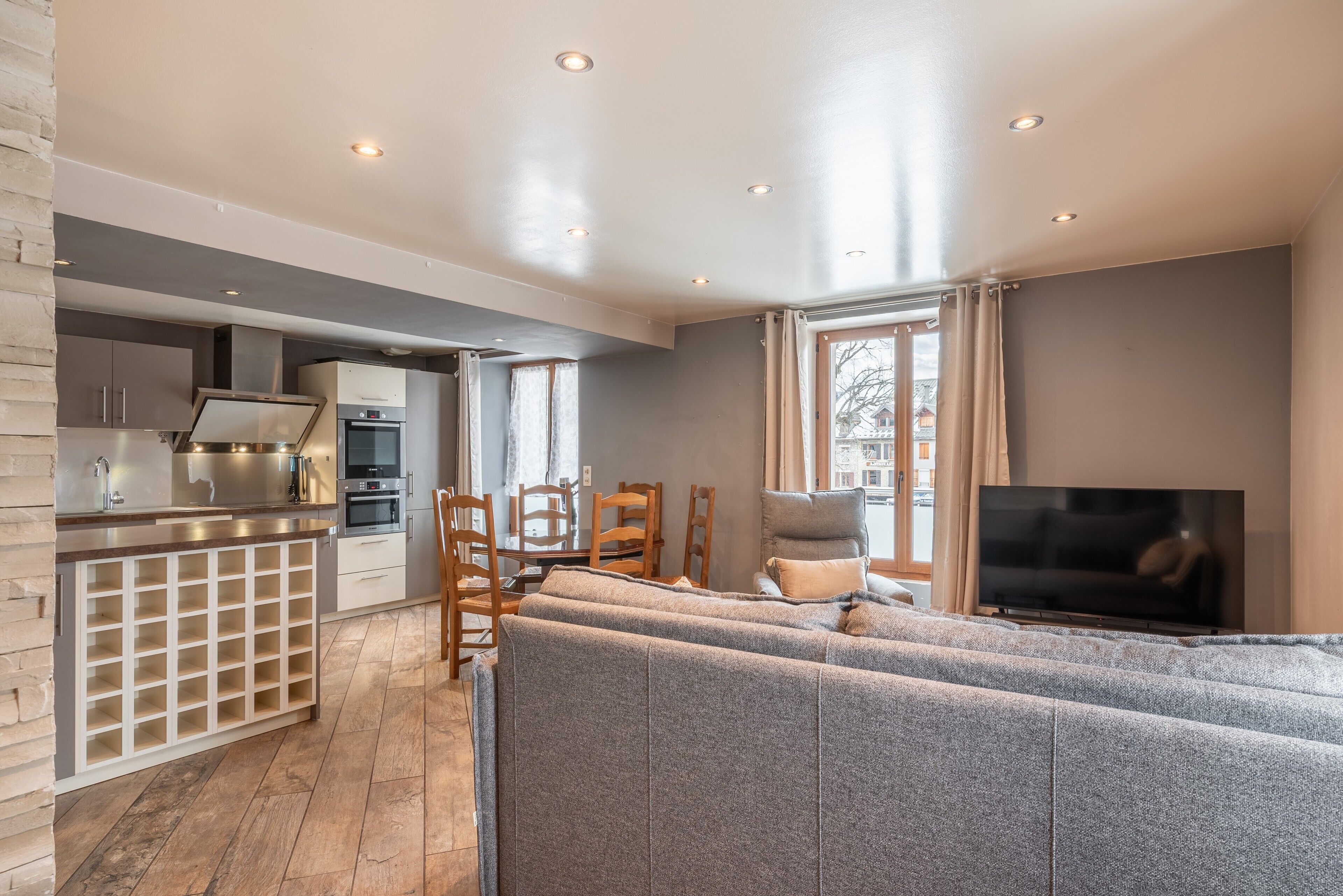 A modern kitchen with built-in appliances, a dining area with wooden chairs, a grey sofa, and a TV.