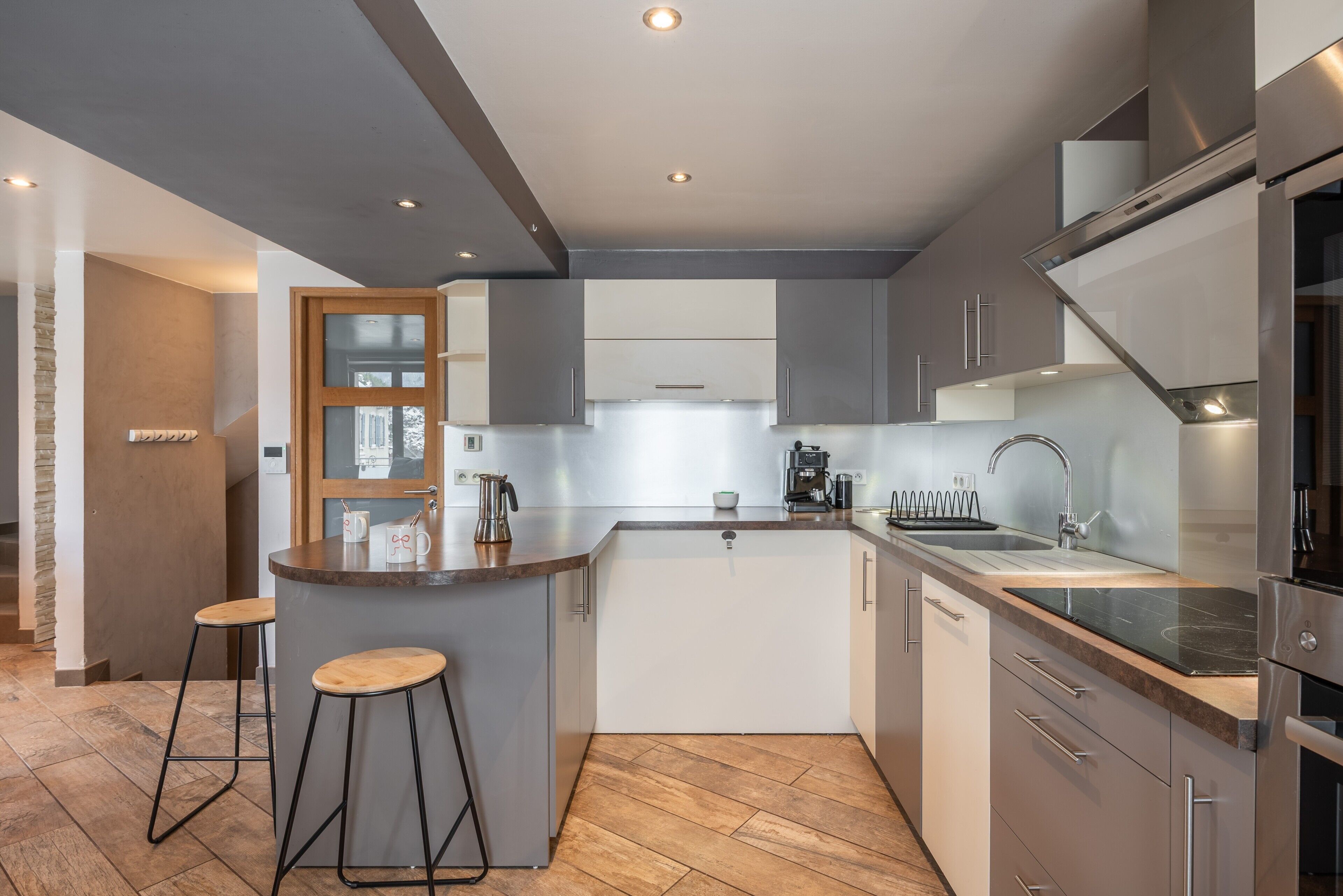 A modern kitchen with a central island, bar stools, stainless steel appliances, and a herringbone floor pattern.