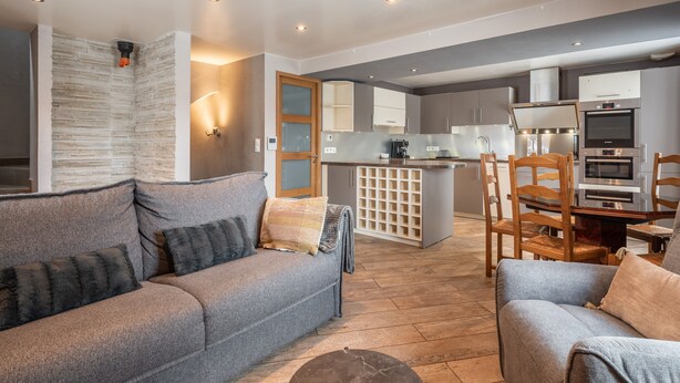 Ein modernes Wohnzimmer mit einem grauen Sofa, Holzstühlen und einer Küche im Hintergrund.