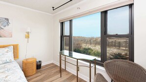 Une chambre avec une grande fenêtre, un lit, une table de chevet, une lampe et un fauteuil en osier.