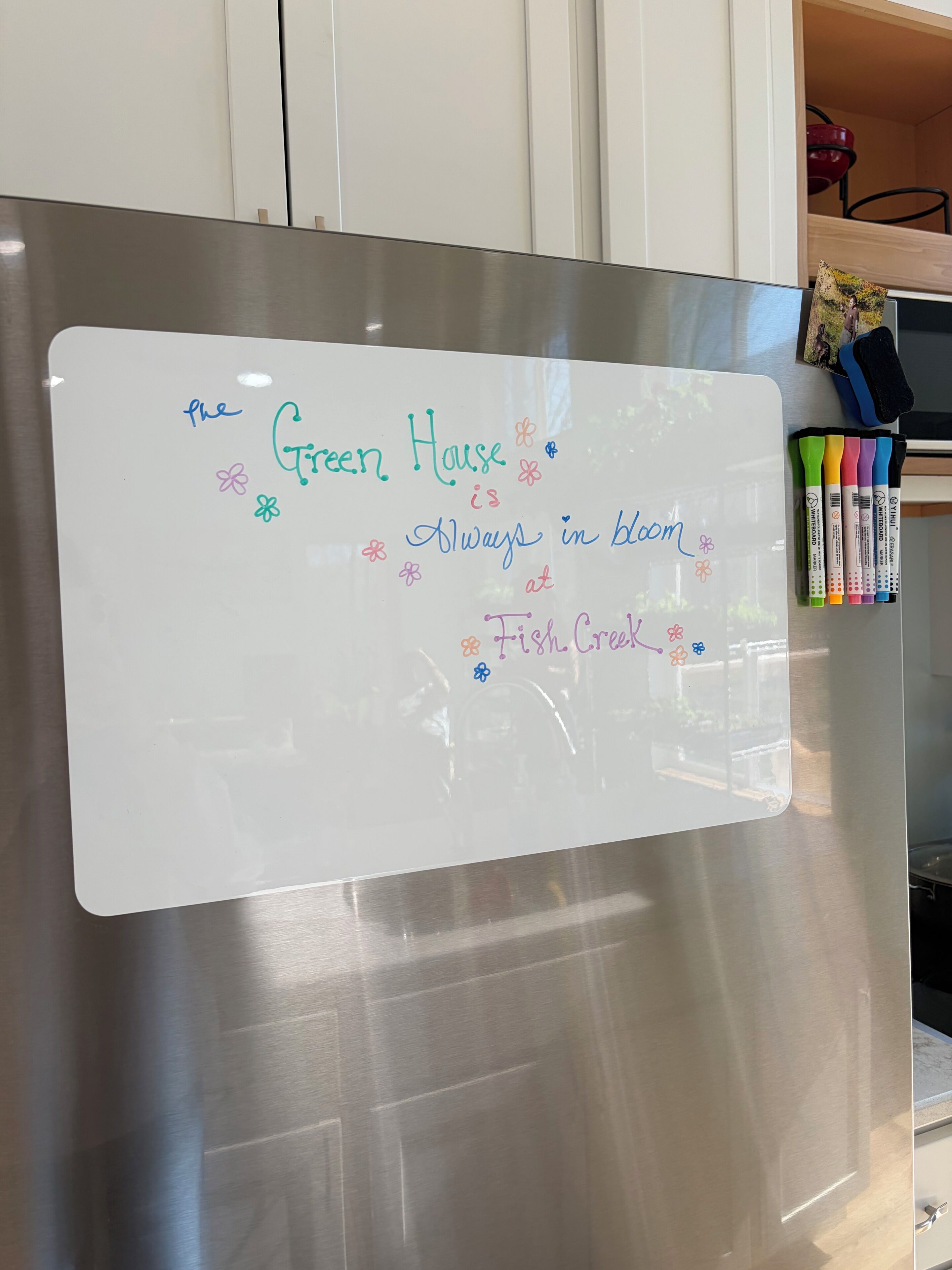 A whiteboard with handwritten text and colorful flowers, located in a kitchen with stainless steel appliances and a wooden cabinet.