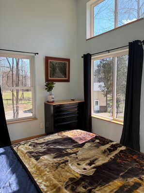 Quarto com cama, escrivaninha com gavetas, quadro emoldurado na parede e grandes janelas com cortinas.