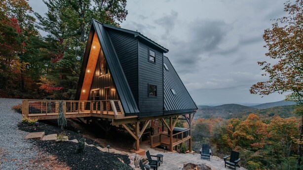 Una cabaña moderna con techo inclinado, terraza de madera, asientos exteriores y un pozo de fuego.