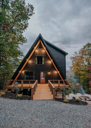 Une cabane avec un toit triangulaire, une terrasse en bois et une allée gravillonnée.