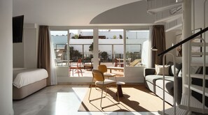 A modern hotel room with a bed, a sofa, a dining area with chairs, a staircase, and a view of the outside.