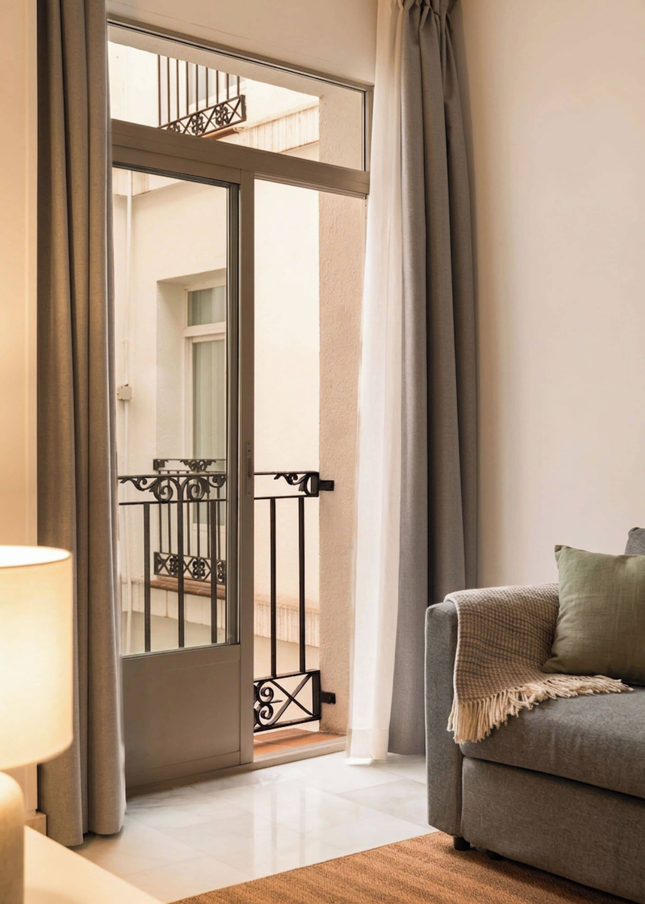 Una habitación con puerta corrediza de vidrio que da a un balcón, un sofá con una manta y una lámpara sobre una mesita auxiliar.