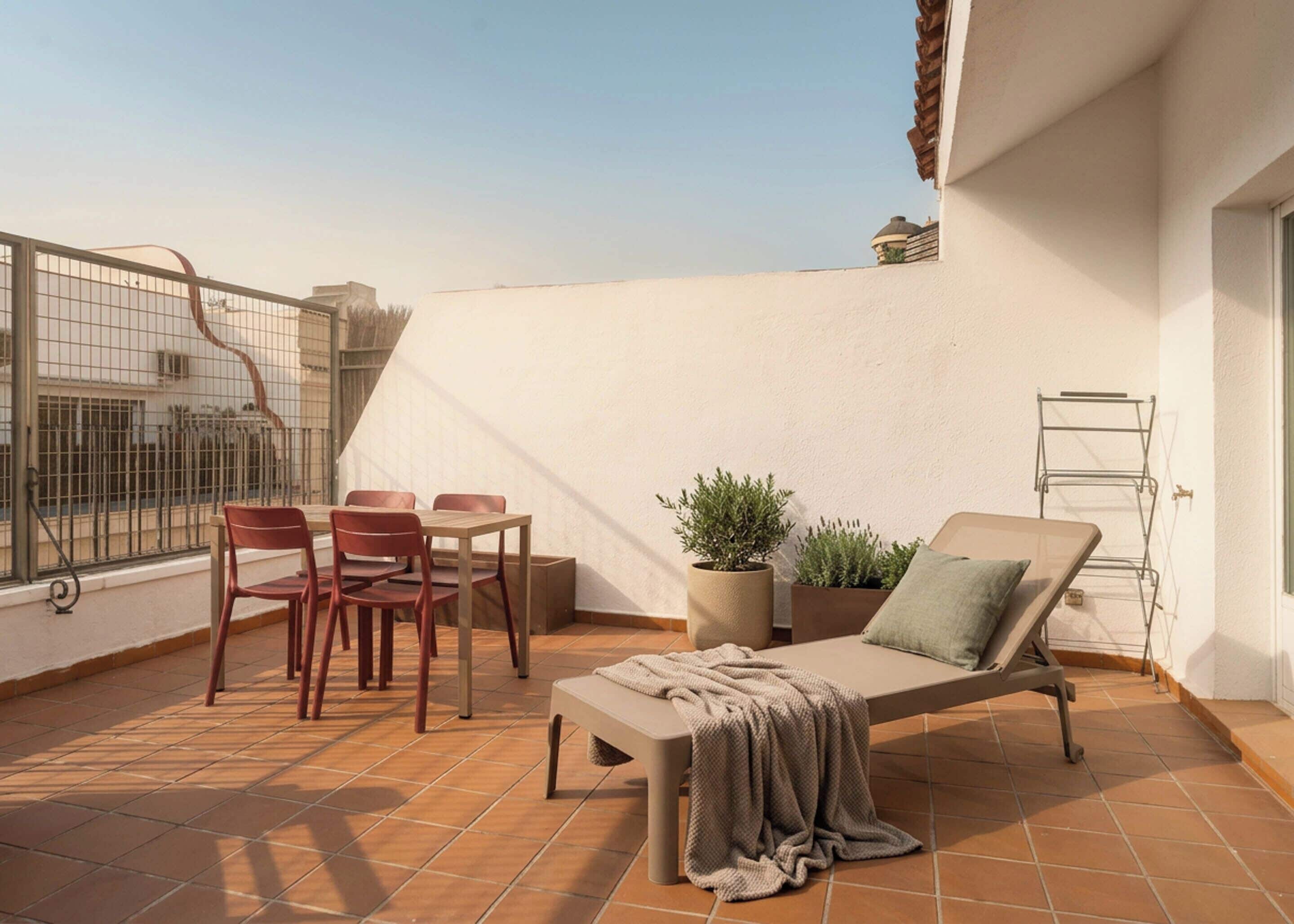 A rooftop terrace with a lounge chair, two chairs, a small table, potted plants, and a metal drying rack.