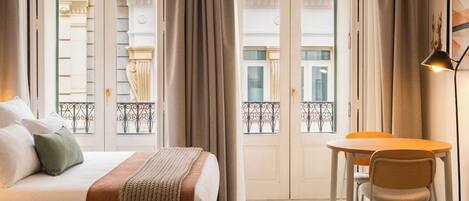 A hotel room with a bed, a desk, and a chair. There are curtains on the windows, and a balcony is visible through the doors.