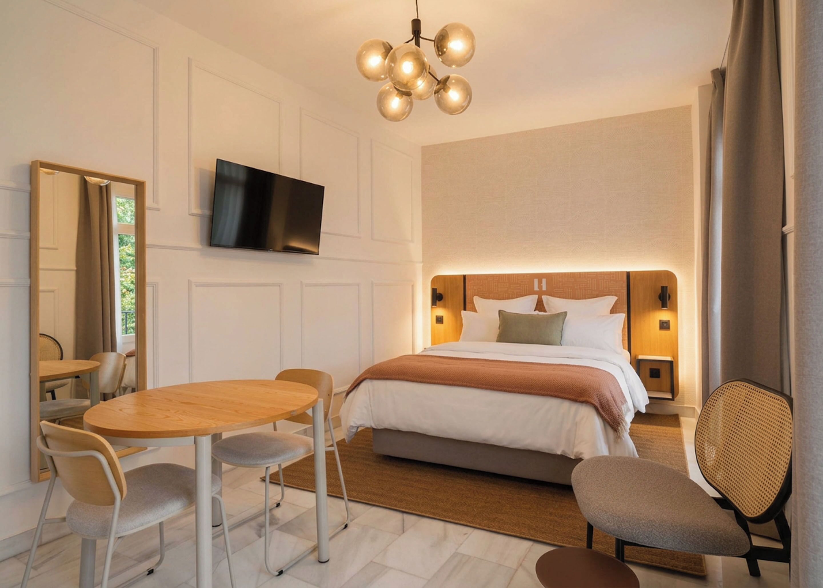 A modern hotel room with a bed, bedside tables, a flat-screen TV, a dining table with chairs, and a decorative ceiling light fixture.