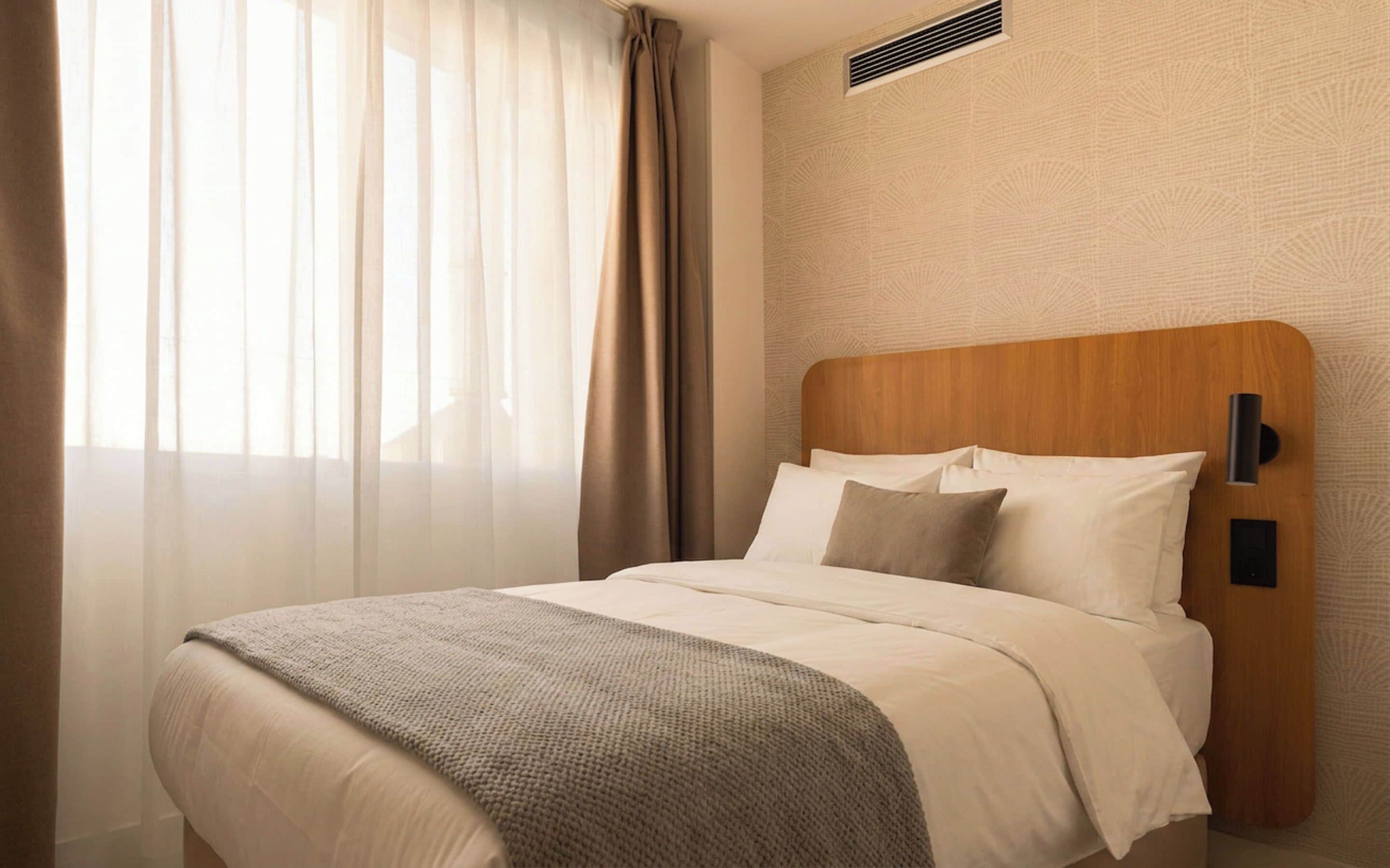 A hotel room with a bed, a wooden headboard, a window with sheer curtains, and a textured wall.