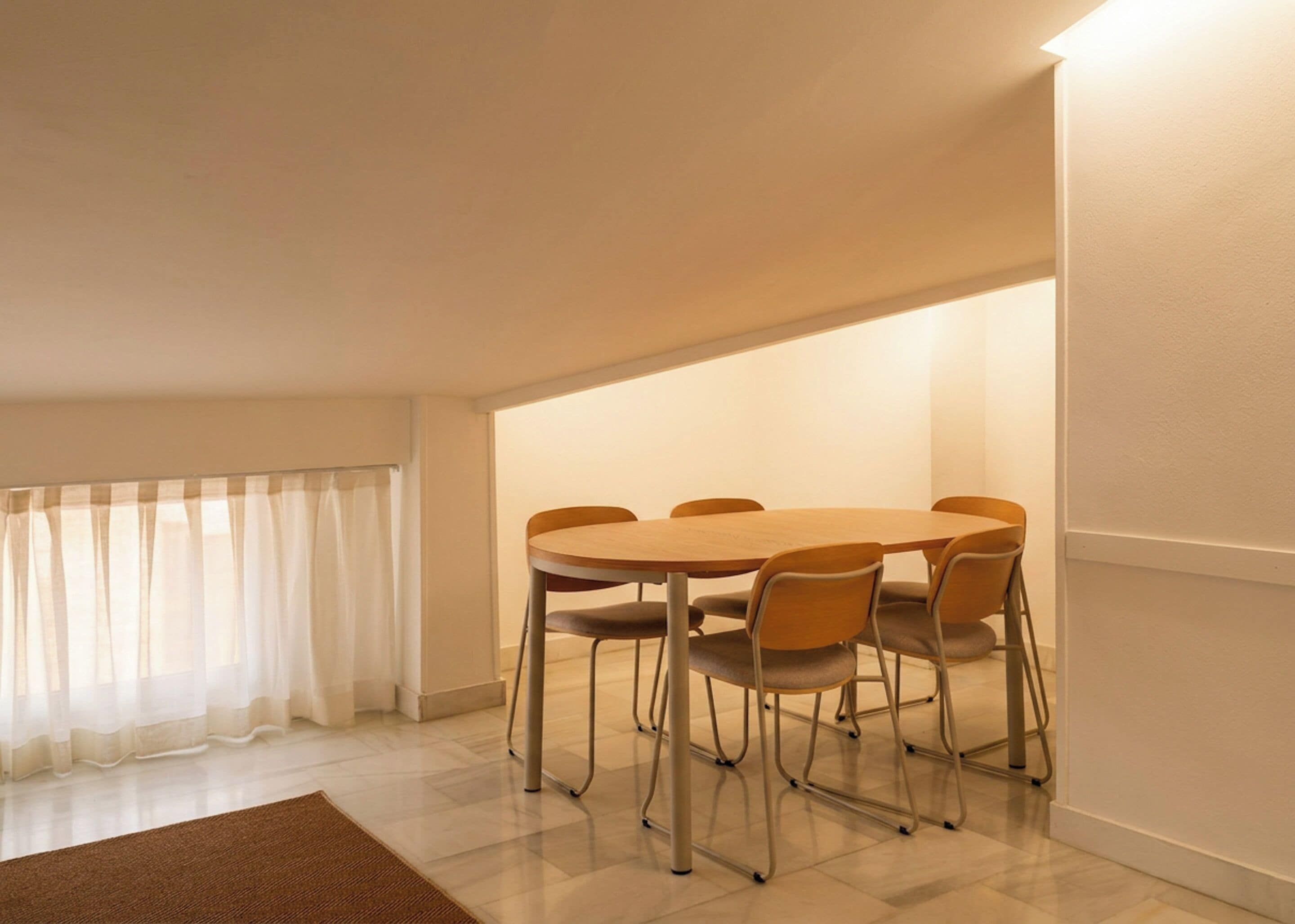A dining area with a round table and six chairs, a window with sheer curtains, and a brown rug on the floor.