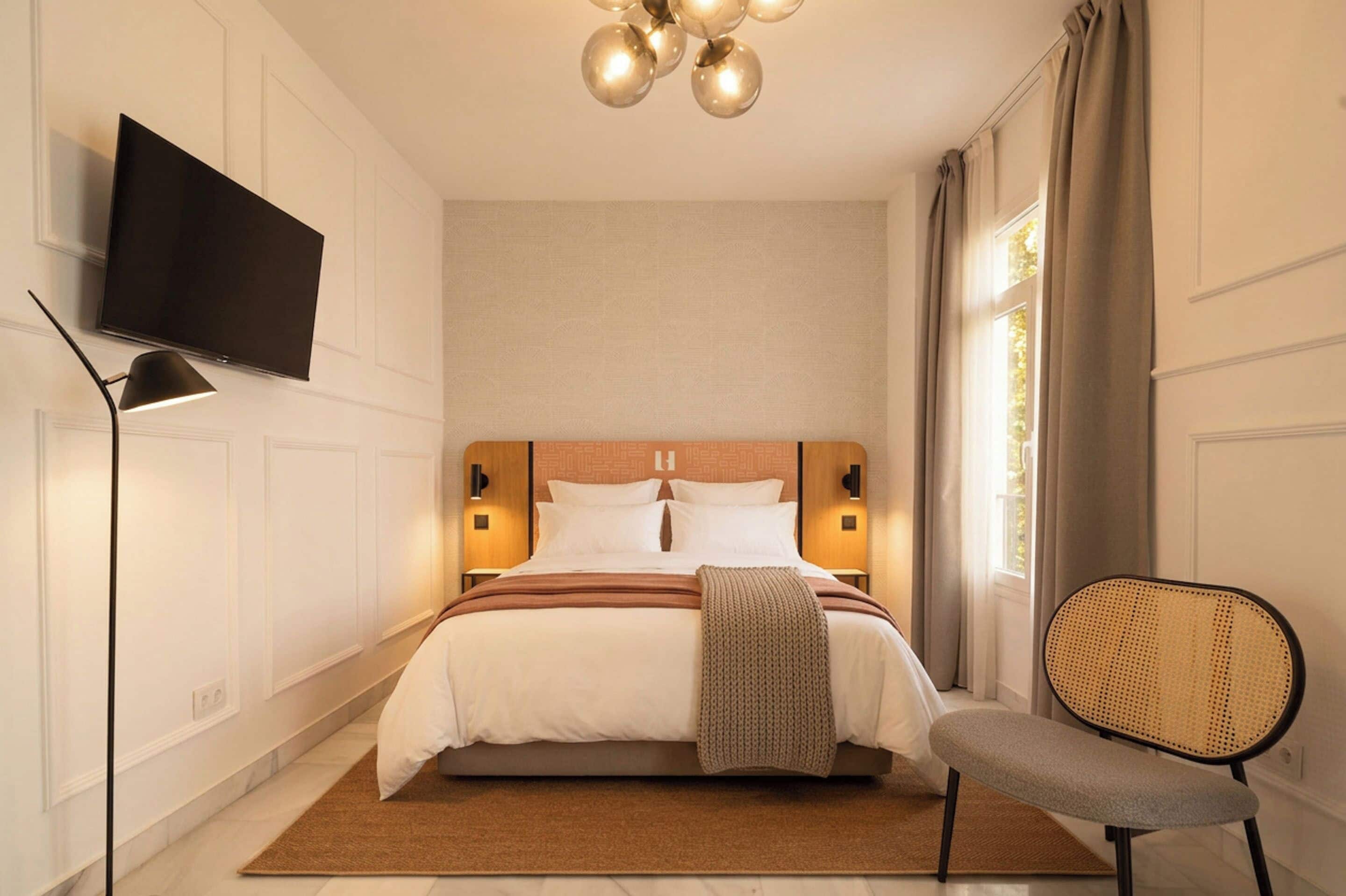 A modern hotel room with a large bed, a flat-screen TV, a bedside lamp, a chair, and a window with curtains.
