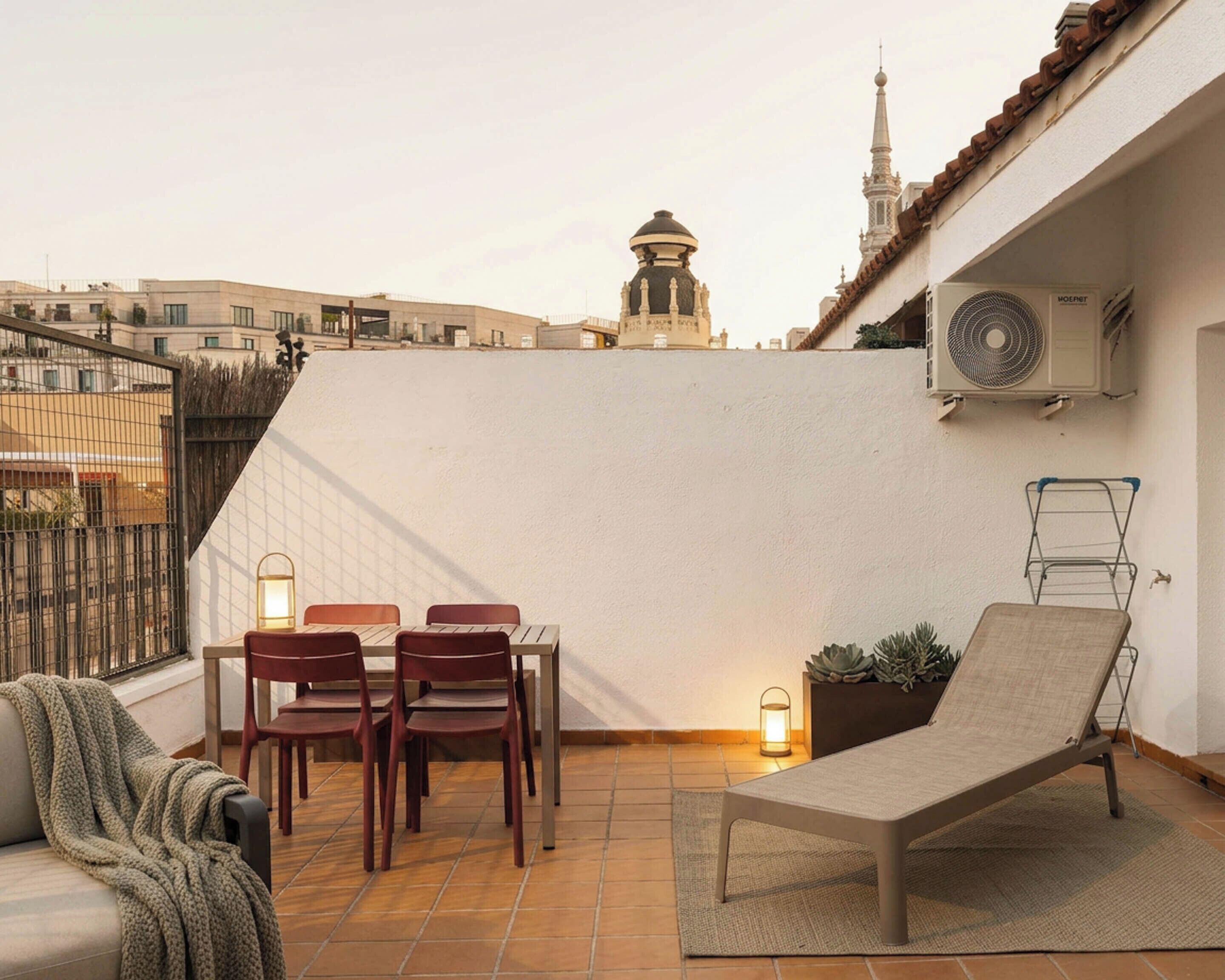 A rooftop terrace with a table and chairs, a lounge chair, a potted plant, and an air conditioning unit.