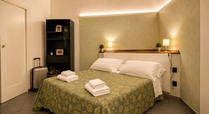 Quarto de hotel com cama, mesa de cabeceira, luminária, prateleira na parede e mala.