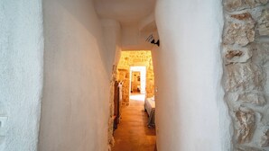 Een smalle gang met witte muren en een stenen muur aan de rechterkant, die leidt naar een kamer met een bed en een houten kledingkast.