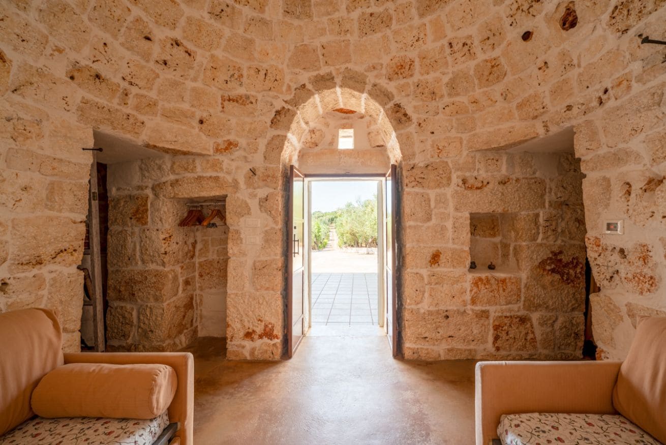 A stone-walled room with an open doorway leading to a courtyard. There are two upholstered chairs with floral upholstery.