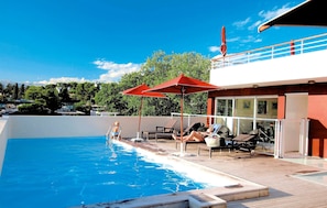 Une piscine avec des chaises longues et des parasols, une terrasse et un bâtiment doté de grandes fenêtres.