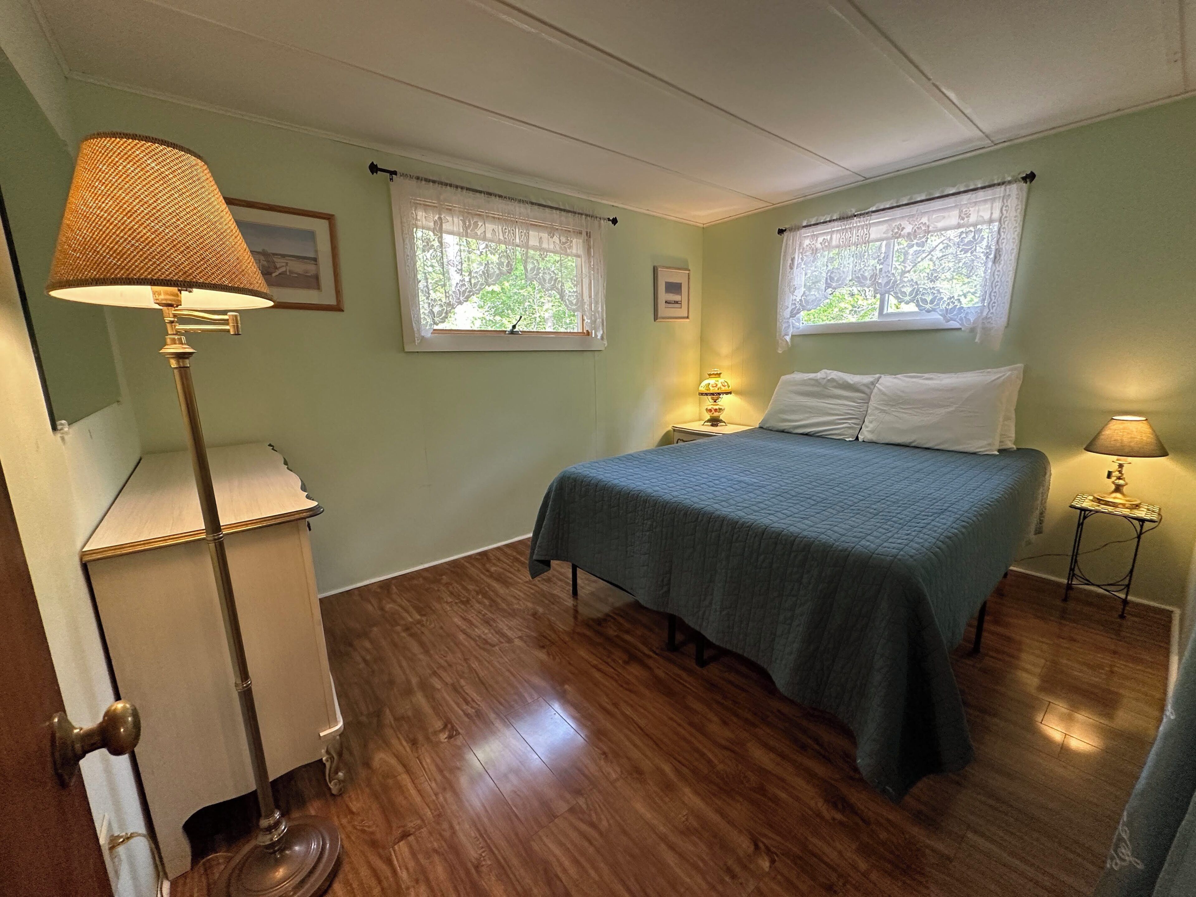Quarto com cama, duas luminárias de cabeceira, uma escrivaninha e duas janelas com cortinas.