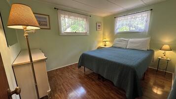 Quarto com cama, duas luminárias de cabeceira, uma escrivaninha e duas janelas com cortinas.