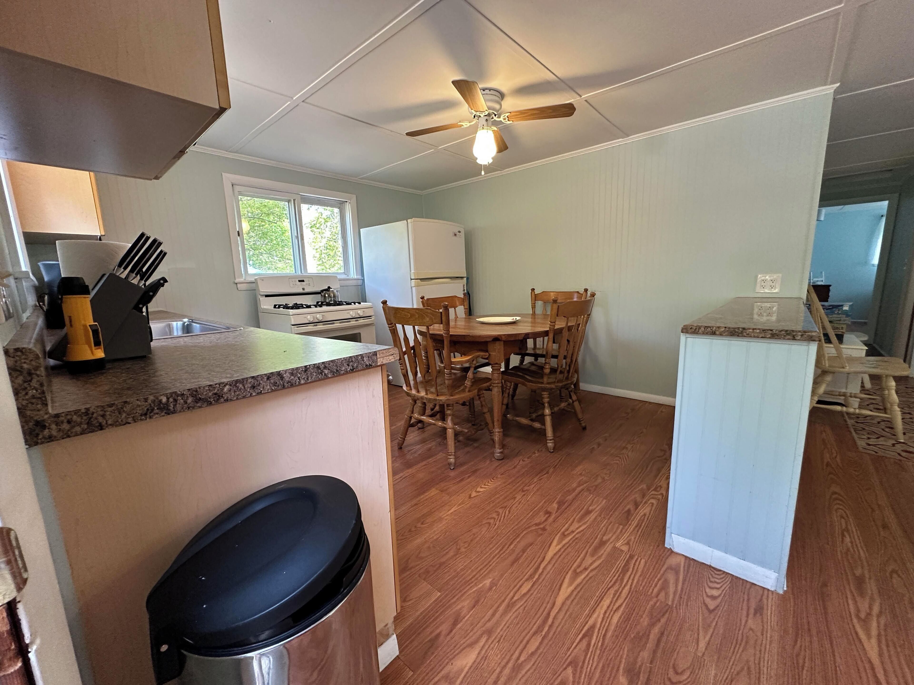 A compact kitchen with a dining area, wooden chairs, a ceiling fan, and a trash bin.