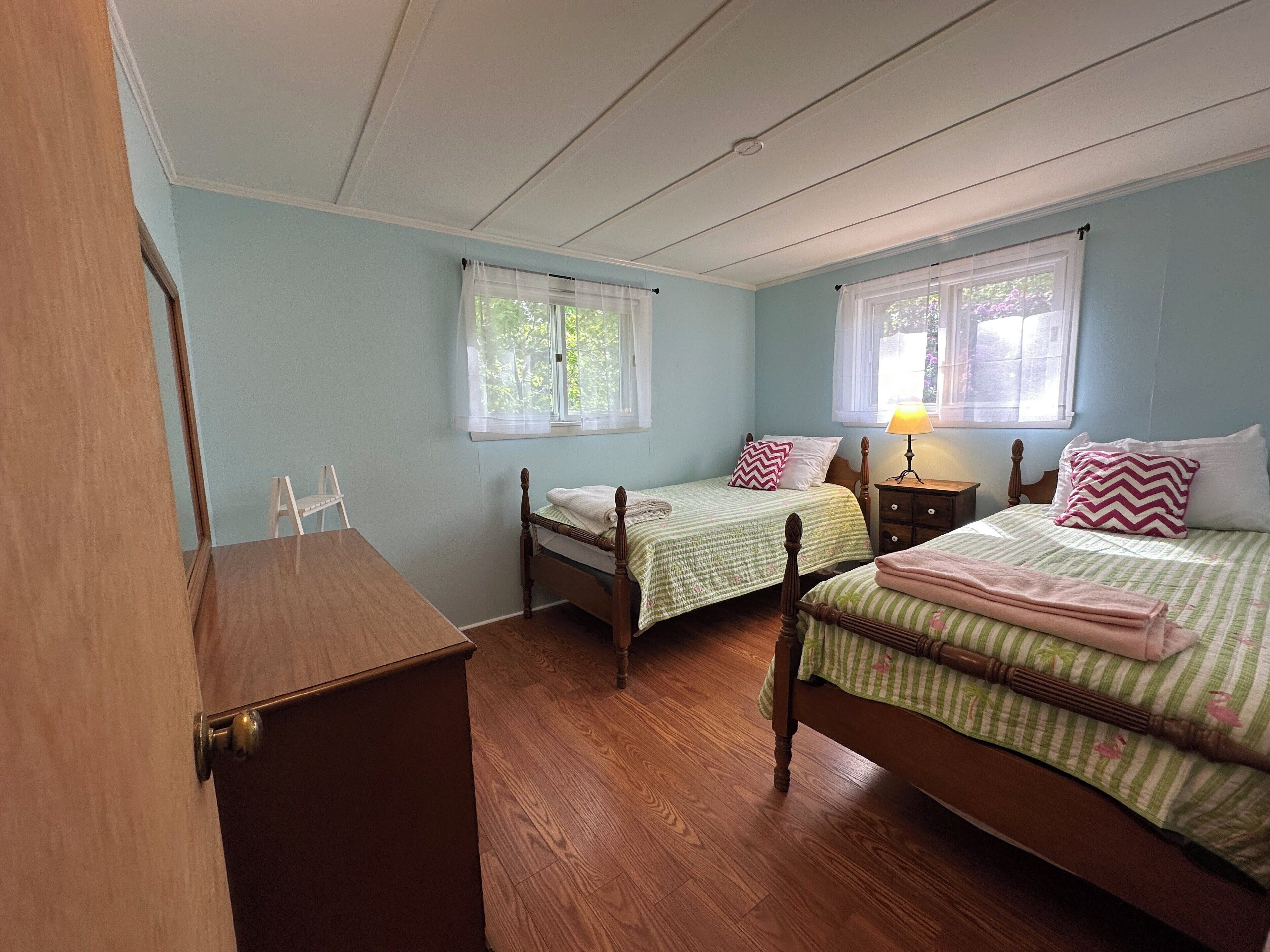 A room with two wooden beds, a wooden dresser, and two windows with sheer curtains.