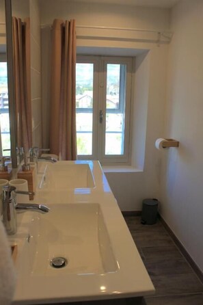 Une salle de bain avec deux lavabos, une fenêtre avec des rideaux, un miroir et une poubelle.