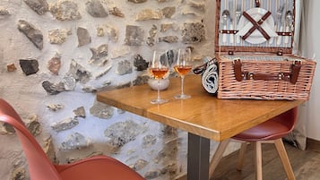 A stone wall interior with a wooden table set for two, two red chairs, and a wicker basket with cutlery.