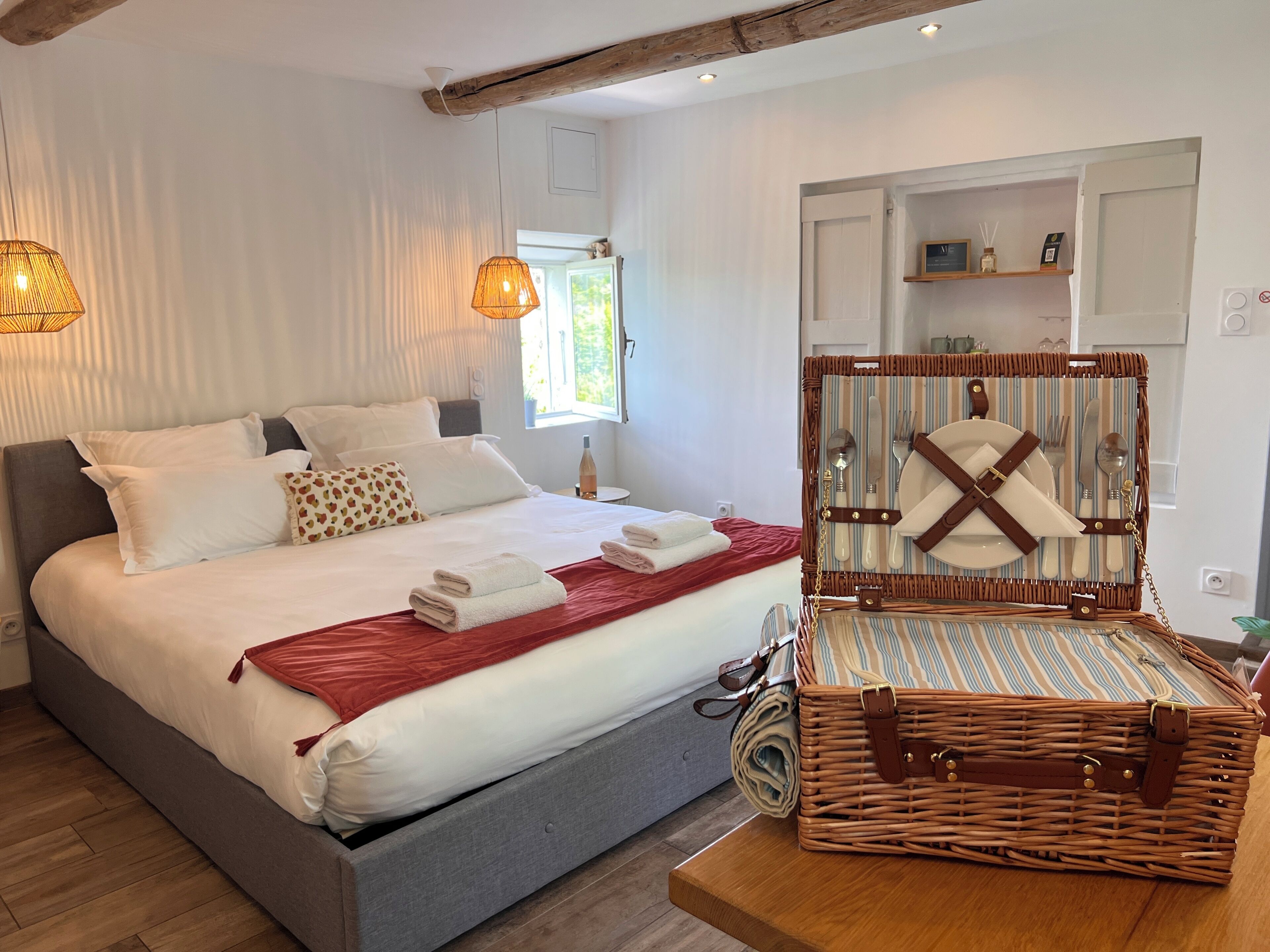 Un lit bien fait, avec du linge de lit blanc et un plaid rouge, une table d’appoint en osier avec une bouteille, et un panier en osier contenant des couverts.