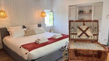 Un lit bien fait, avec du linge de lit blanc et un plaid rouge, une table d’appoint en osier avec une bouteille, et un panier en osier contenant des couverts.