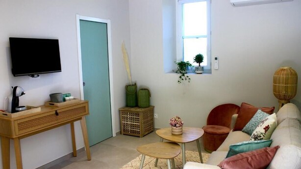 Un salon comprenant un téléviseur, un bureau en bois, un canapé avec des coussins, une petite table et une fenêtre avec des plantes.