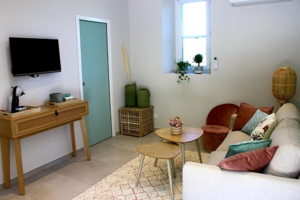 Un salon comprenant un téléviseur, un bureau en bois, un canapé avec des coussins, une petite table et une fenêtre avec des plantes.