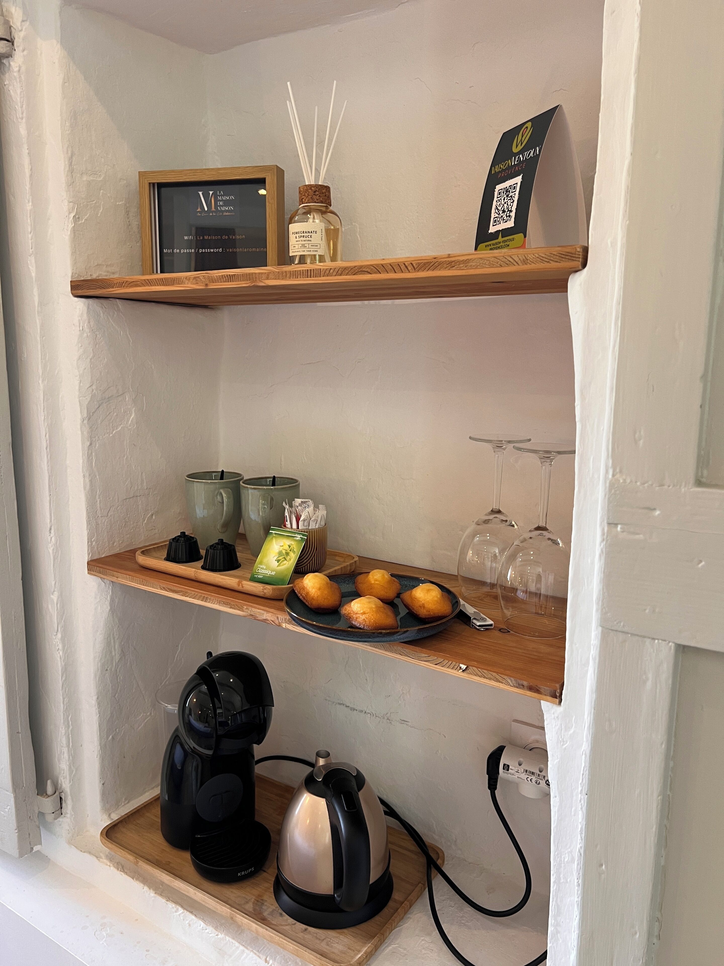 Une étagère avec un panneau encadré, un diffuseur d’arômes et un code QR. En dessous, on trouve des théières, un plateau avec des pâtisseries et une bouilloire.