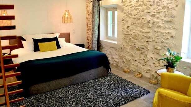 Ein Schlafzimmer mit Bett, Fenster, einer Steinwand, einer Pflanze und einem gelben Stuhl.