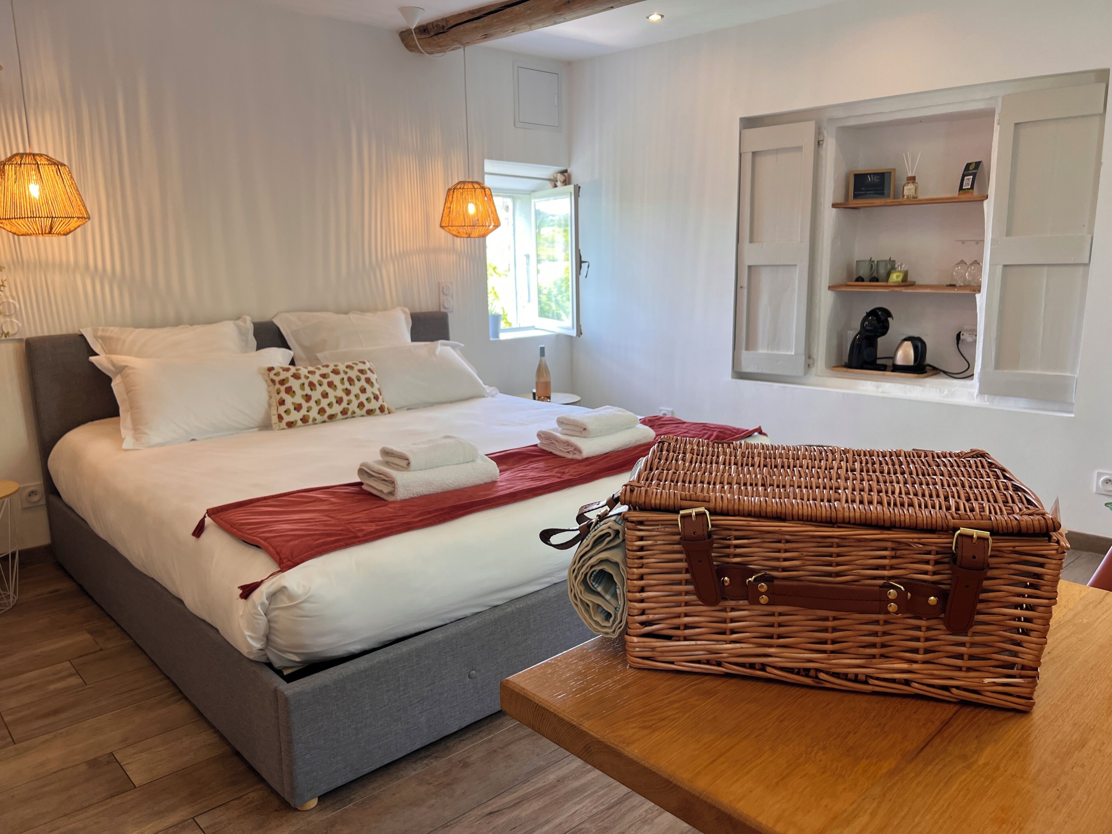 Un lit bien fait, avec du linge de lit blanc et un plaid rouge, un panier en osier posé sur une table en bois, et une fenêtre donnant sur un paysage verdoyant.