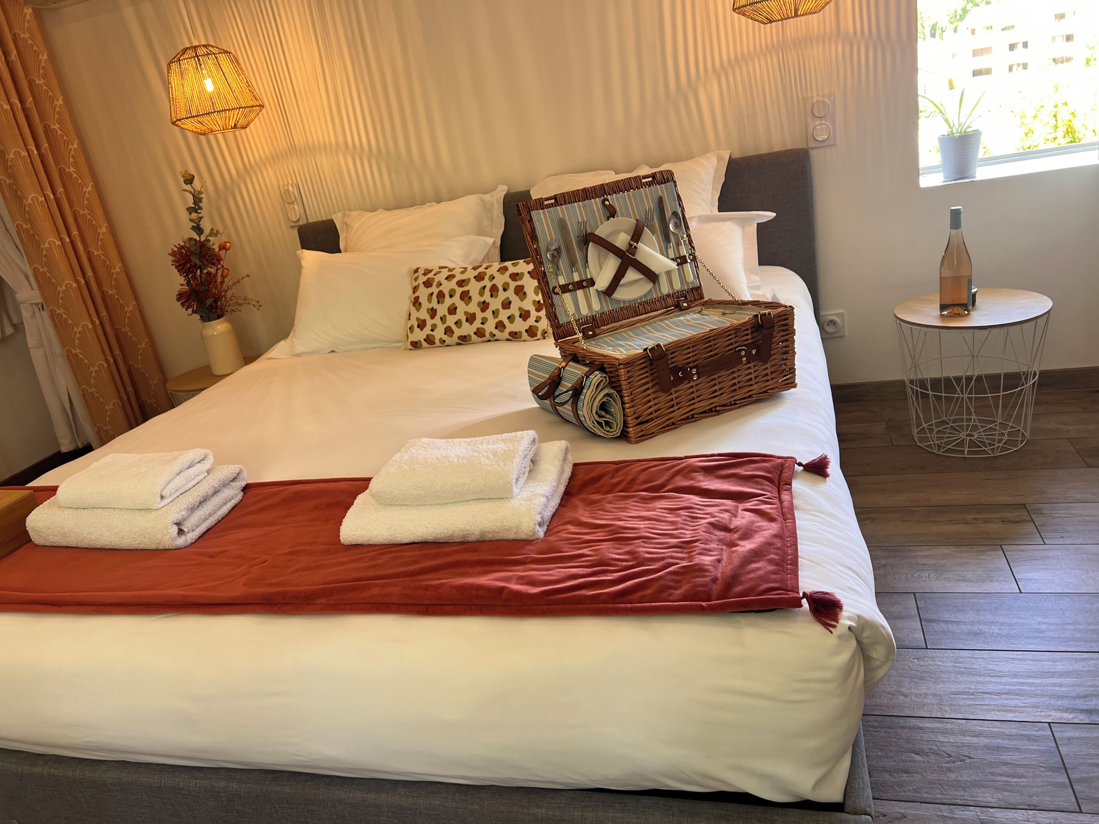 Una cama bien hecha con sábanas blancas, una manta roja y una maleta de mimbre sobre la cama.