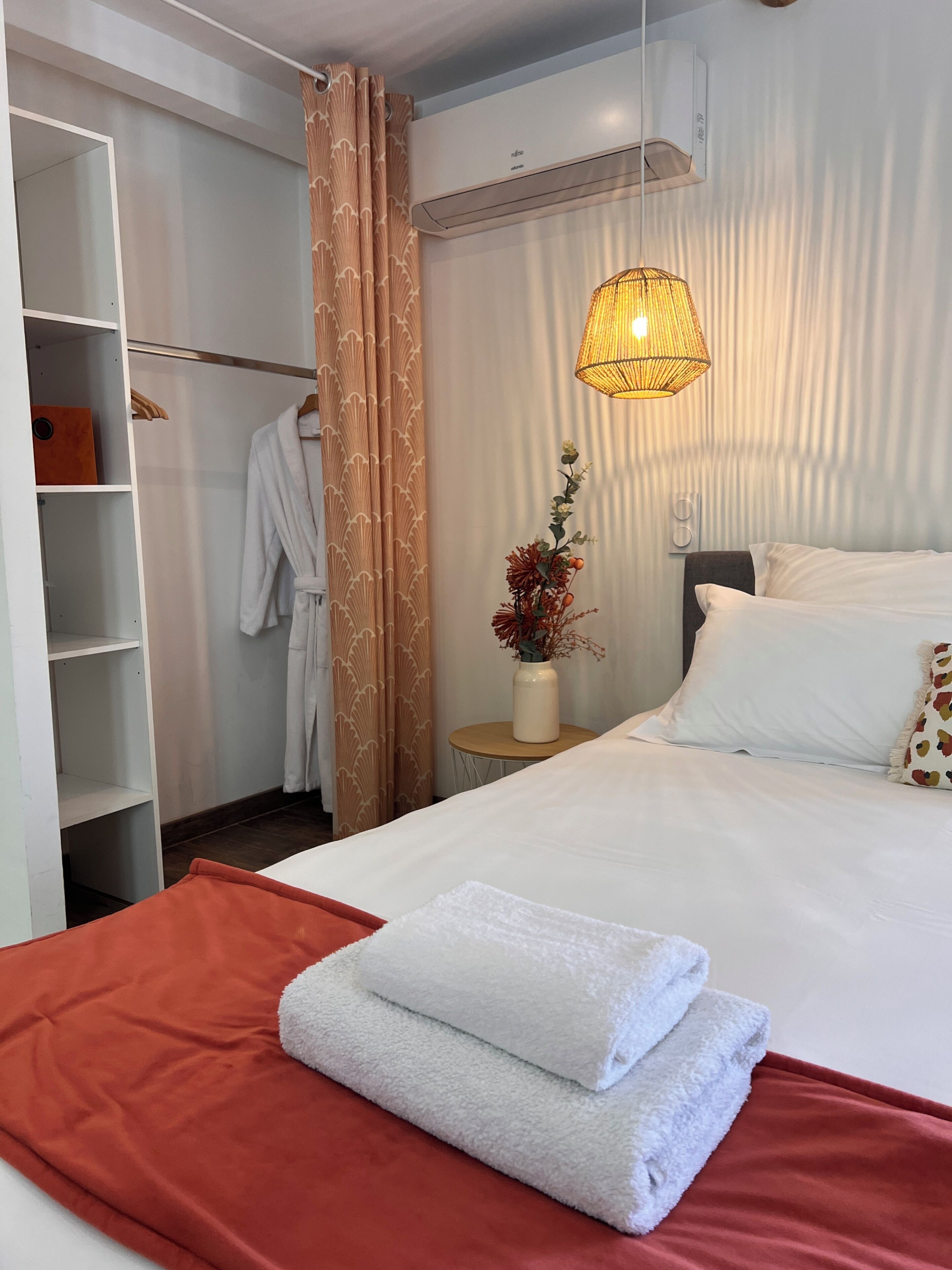 A hotel room with a bed, a wardrobe, a hanging robe, a vase with flowers, and a wall-mounted air conditioner.